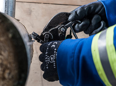 A person wearing black gloves and a blue jacket with reflective stripes is demonstrating reliability engineering skills by using a pipe cutter to efficiently cut through a metal pipe. The background is a wooden surface, emphasizing the precision and professionalism involved in such services.