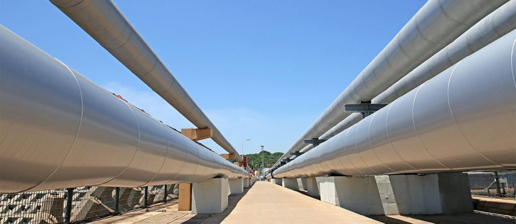 Close-up view of large, parallel steel pipelines stretching into the distance under a clear blue sky, their integrity assured by regular inspection services. The pipes are mounted on concrete supports, with greenery visible at the far end.
